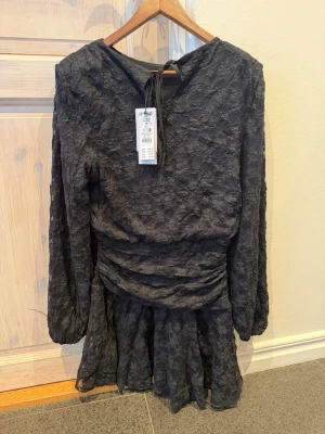 Svart spetsklänning med volang - Säljer en svart kort klänning i spets med lång ärm och volang nedtill. Klänningen har en omlottliknande midja och knytband i nacken. Perfekt för dig som gillar romantiska detaljer och en feminin siluett.