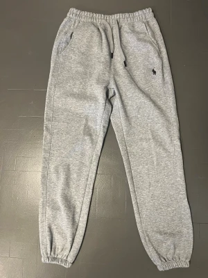 Grå mjukisbyxor från Polo Ralph Lauren - Säljer ett par grå mjukisbyxor från Polo Ralph Lauren med elastisk midja och snörning. Byxorna har sidofickor och den klassiska Polo-loggan broderad på vänster sida. Materialet är mjukt och skönt. Passformen är loose och muddar vid benslut. Även om de såklart passar på någon med storlek M tror jag att de hade passat bättre på någon med S eller till och med XS. QR koden fungerar.