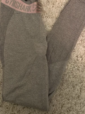 Grå leggings från Gymshark - Snygga grå leggings från Gymshark med bred resår i midjan och logga framtill. De är tillverkade i ett stretchigt material som sitter tajt och har diskreta sömdetaljer för en sportig look. Perfekta för träning eller chill.