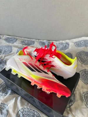 Adidas F50  - Snygga f50 fotbollsskor från Adidas i vitt med röda och gula detaljer samt svarta ränder på sidan. Skorna har snörning och en låg profil, perfekt för snabba rörelser på planen. Det är storlek 43 och en tredjedel!! Yttermaterialet är syntet och sulan har dobbar för bra grepp på gräs. För fler bilder skriv till mig!✌️