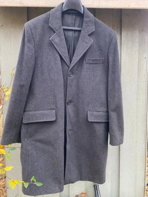 Mörkgrå ullkappa med klassisk krage - Stilren mörkgrå kappa i ullmix med klassisk krage och två knappar framtill. Kappan har två stora fickor med lock och en bröstficka. Lång modell som passar perfekt till höst och vinter. Fodrad insida för extra komfort.