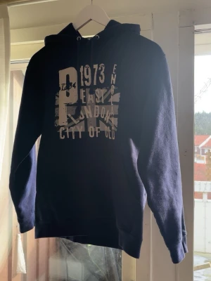Mörkblå hoodie med tryck - Snygg mörkblå hoodie med stor vit och grå print framtill som visar texten '1973 East London City of Old England' och en brittisk flagga i bakgrunden. Hoodien har dragsko i huvan och ribbade muddar vid ärmar och midja. Perfekt för en avslappnad streetwear-look. Passar i xs va för liten när jag köpte den inte så mycket använd