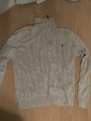 Beige kabelstickad tröja Polo Ralph Lauren - Säljer en grä kabelstickad tröja från Polo Ralph Lauren med halv dragkedja och hög krage. Tröjan har det klassiska broderade logotypen på bröstet och ribbade muddar vid ärmslut och nederkant. Perfekt för lager på lager och snygg till jeans.