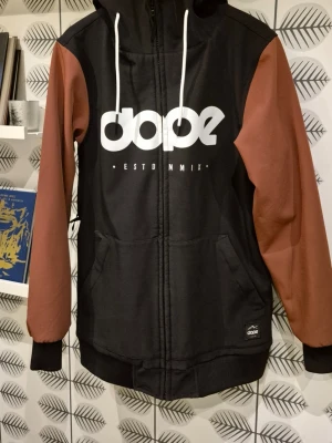 Skid/snowboardjacka Dope, svarta skidbyxor på köpet - Dope skidjacka, svart och brun, i mycket gott skick. Jackan har huva med vita snören, dragkedja framtill och två stora fickor, innerficka och ficka för liftkort i ärmen. Ribbade muddar vid ärmslut och nederkant. Skickar med ett par svarta Dope skidbyxor, också dessa i stl S, i ok skick men lite slitna i färgen på knän och rumpa.