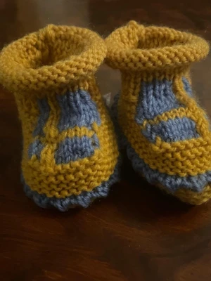 Stickade tofflor i gult och blått - Handstickade tofflor i mjukt garn med gul bas och blå detaljer. Perfekta för att hålla fötterna varma och mysiga. Snyggt mönster och stretchig passform som gör dem bekväma att bära.