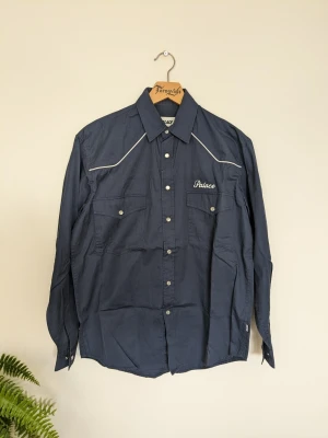 Palace Western Skjorta - Snygg mörkblå "western" skjorta från Palace med vita kontrastsömmar och broderad logga på bröstet. På ryggen står det 'Palazzo Londra' i stor vit brodyr. Skjortan har knappar framtill och två bröstfickor med lock.