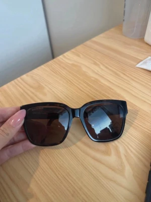 Bruna solglasögon från Balenciaga - Snygga bruna solglasögon från Balenciaga med bred båge och mörka fyrkantiga glas. Klassisk och trendig design med logga på insidan av skalmen. Perfekta för att lyfta din stil till nästa nivå.