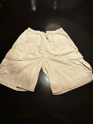 Beige cargoshorts med snörning - Säljer ett par beige cargoshorts från Cava Company med flera praktiska fickor på sidorna och snörning i midjan. Shortsen har en avslappnad passform och är perfekta för varma dagar. Klassisk cargostil med elastisk midja för extra komfort.