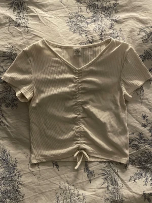 Vit ribbad croppad topp med knyt - Säljer en ljusvit ribbad topp med v-ringning och korta ärmar. Toppen är croppad och har en snygg snörning med rosett framtill som ger en trendig look. Perfekt att matcha med jeans eller kjol.