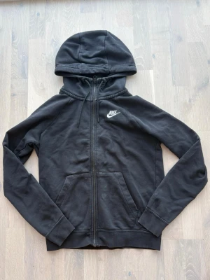 Svart zip hoodie från Nike - Klassisk svart hoodie från Nike med dragkedja och vit broderad logo på bröstet. Hoodien har justerbar huva med snörning, kängurufickor och ribbade muddar. Perfekt till chill eller streetwear. Mjuk bomullsblandning för skön känsla.