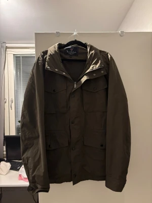 Olivgrön vindjacka från Brandblack - Snygg olivgrön vindjacka från Brandblack med fyra stora fickor framtill, dragkedja och tryckknappar. Jackan har hög krage och justerbar huva. Perfekt för blåsiga dagar och enkel att matcha med olika outfits.