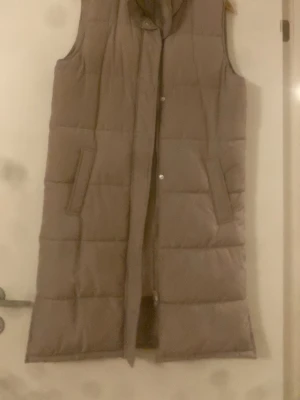 Lång beige dunväst från Levete Room - Stilren lång beige dunväst från Levete Room med quiltad design och två snedställda fickor. Västen har hög krage och stängs med tryckknappar. Perfekt för lager på lager och ger en trendig look till din outfit.