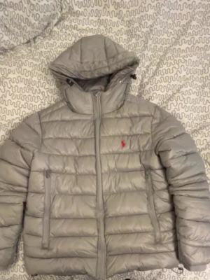 Grå pufferjacka från Polo Ralph Lauren - Snygg grå pufferjacka från Polo Ralph Lauren med huva och dragkedja framtill. Jackan har två sidofickor med dragkedja och den klassiska röda Polo-loggan på bröstet. Tillverkad i lätt, quiltat material som håller värmen under kalla dagar.