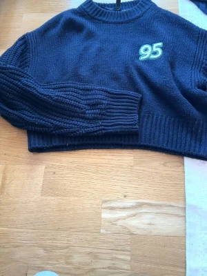 Mörkblå cropped stickad tröja - Säljer en mörkblå cropped stickad tröja med broderad siffra '95' i ljusblått och vitt på bröstet. Tröjan har ribbstickade muddar och en klassisk rund halsringning. Perfekt för en avslappnad och trendig look.