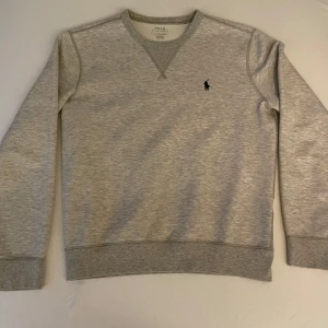 Grå sweatshirt från Ralph Lauren - Klassisk ljusgrå sweatshirt från Ralph Lauren med rund halsringning och ribbade muddar. Tröjan har den ikoniska svarta Polo-loggan broderad på bröstet. Tillverkad i mjuk bomullsblandning för en skön och avslappnad look.