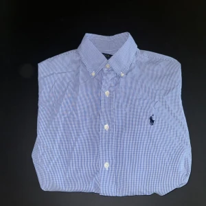 Blårutig Oxford skjorta från Polo Ralph Lauren - Klassisk blårutig skjorta från Polo Ralph Lauren med button-down krage och broderad logga på bröstet. Skjortan har lång ärm och knäppning framtill. Perfekt för en stilren och tidlös look. Skjortan är slim. 