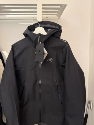 Svart vindjacka från Arc'teryx - Snygg svart vindjacka från Arc'teryx med huva och dragkedja framtill. Aldrig använd