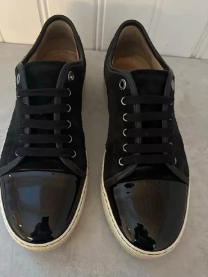 Svarta Lanvin sneakers storlek 41 - Snygga svarta sneakers från Lanvin med klassisk låg profil. Skorna har ovandel i mocka och en glansig lackad tå, vita sulor och svarta skosnören. Perfekta för dig som gillar stilrena och exklusiva sneakers med en twist.