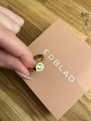 Guldfärgad ring med sten från Edblad - Snygg guldfärgad ring från Edblad med en rund, klar sten i mitten. Ringen har en modern och stilren design som passar perfekt för dig som gillar minimalistiska accessoarer. Materialet ser ut att vara metall med en polerad finish.
