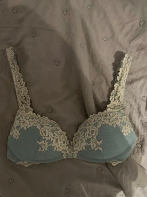 Ljusblå spets-bh med blommönster - Supersnygg ljusblå bh med vadderade kupor och dekorativt blommönstrad spets i beige längs kanterna och axelbanden. Bh:n har justerbara axelband och hakknäppning bak. Perfekt för dig som gillar romantiska detaljer och mjuka pastellfärger.