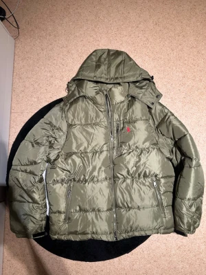 Olivgrön pufferjacka från Polo Ralph Lauren - Säljer en olivgrön pufferjacka från Polo Ralph Lauren med avtagbar huva och tvåvägsdragkedja. Jackan har två framfickor med dragkedja och den klassiska röda Polo-loggan broderad på bröstet. Materialet är glansigt och quiltat för extra värme.