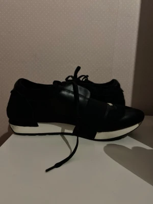 Svarta sneakers med vit sula - Snygga svarta sneakers med vit sula och svarta skosnören. Skorna har en modern design med blandning av läder och textil samt en bred elastisk rem över foten för extra stil. Perfekta för dig som gillar en clean och sportig look.