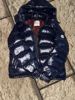 Mörkblå glansig dunjacka Moncler - Snygg mörkblå dunjacka från Moncler med glansigt yttertyg och vinrött foder. Jackan har huva, dragkedja framtill och Moncler-logga på ärmen. Perfekt för kalla vinterdagar och riktigt trendig look.