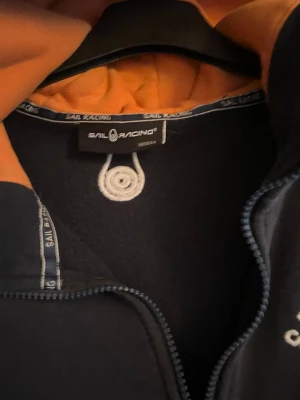 Marinblå hoodie från Sail Racing - Snygg marinblå hoodie från Sail Racing med dragkedja och orangefärgat foder i huvan. Hoodien har detaljerade logotexter på insidan och en liten logopatch vid fickan. Perfekt för dig som gillar sportig och stilren design.
