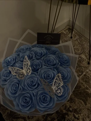 Blå rosbox med dekorfjärilar Eternelle - Säljer en bukett med handgjorda blå rosor, dekorerad med två vita dekorationsfjärilar. Buketten är inslagen i transparent papper och har ett elegant utseende, perfekt som present eller dekoration. Märket Eternelle syns på ett kort i buketten.