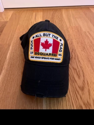 Svart Dsquared2 keps med patch - Säljer en svart keps från Dsquared2 med stor broderad patch framtill med kanadensisk lönnlöv och text. Kepsen har böjd skärm och justerbar rem bak. Materialet är bomull och detaljerna är i rött, vitt och gult. Perfekt för dig som vill sticka ut med en statement-accessoar.