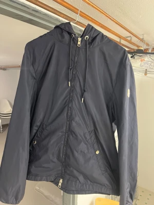 Mörkblå vindjacka från Moncler - Snygg mörkblå vindjacka från Moncler med huva och dragsko. Jackan har två fickor med knappstängning och dragkedja framtill. Moncler-logga på ärmen ger en exklusiv touch. Perfekt för blåsiga dagar och enkel att matcha med olika outfits.