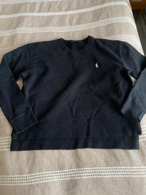 Svart sweatshirt från Ralph Lauren - Klassisk svart sweatshirt från Ralph Lauren med den ikoniska vita polospelaren broderad på bröstet. Tröjan har rund halsringning, ribbade muddar och är tillverkad i mjukt bomullsmaterial. Perfekt för en avslappnad och stilren look. 