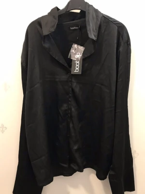 Svart satinset från Boohoo - Snyggt svart set från Boohoo i mjukt satinmaterial. Setet består av en långärmad skjorta med krage och knappar framtill samt matchande vida byxor med resår i midjan. Perfekt för dig som gillar en stilren och trendig look.