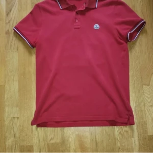 Röd pikétröja från Moncler - Säljer en klassisk röd pikétröja från Moncler med vit och marinblå rand på krage och ärmslut. Tröjan har knappar framtill och Moncler-logga på bröstet. Perfekt för en stilren och sportig look.