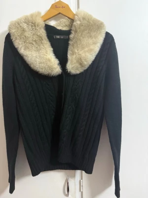 Svart kofta med fuskpäls-krage - Snygg svart stickad kofta med bred, mjuk fuskpäls-krage i beige. Koftan har kabelstickat mönster framtill, långa ärmar och ribbade muddar. Perfekt för dig som vill ha en unik och mysig look med vintage-vibbar.