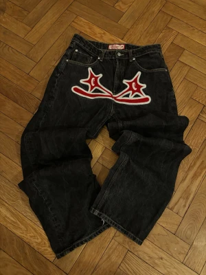 Svarta jeans från Protect london - Unika svarta jeans från Protect london med bred passform och stor röd-vit brodyr framtill. Jeansen har klassisk femficksdesign och cool streetwear-vibe.