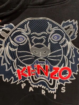 Svart Kenzo hoodie  - Svart hoodie från Kenzo med ikoniskt tigertryck i blått och vitt samt röd logga på bröstet. Hoodien har huva med dragsko och stor magficka framtill. Materialet är mjukt och skönt, 