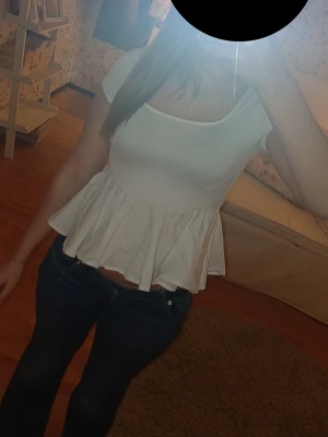 Vit peplum topp med öppen rygg - Snygg vit peplum topp med korta ärmar och rund halsringning. Toppen har en volang nertill och öppen rygg med knytband, vilket ger en trendig och feminin look. Perfekt att styla med jeans eller kjol för en fräsch outfit.