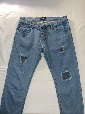 Ljusa slitna jeans från Dsquared2 - Säljer ett par ljusblå jeans från Dsquared2 med flera slitningar och hål på benen för en trendig, avslappnad look. Klassisk femficksmodell med raka ben och synliga sömmar. Perfekta för dig som gillar streetstyle och vill sticka ut.