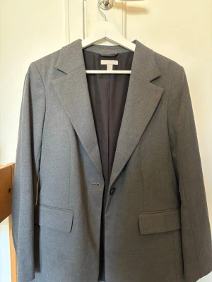 Grå kavaj från H&M med klassisk passform - Stilren grå kavaj från H&M med klassisk passform och enkelknäppning. Kavajen har två fickor med lock framtill och är helfodrad. Använt endast en gång. Den är något oversize i storleken