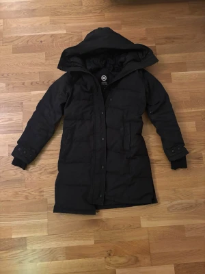 Äkta Svart dunjacka från Canada Goose - Svart lång dunjacka från Canada Goose med huva och klassisk logga på ärmen. Jackan har knappar och dragkedja framtill samt ribbade muddar vid ärmsluten. Perfekt för kalla vinterdagar och riktigt varm tack vare dunfyllningen. Canada goose påsen kommer även med. Jackan är även äkta och bevis finns på bild 6.