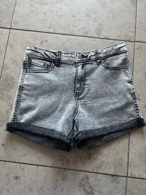 Grå denimshorts från Cubus - Snygga grå denimshorts från Cubus med klassisk femficksdesign och uppvikta benslut. Shortsen har knapp och dragkedja framtill samt tydliga svarta sömmar som ger en cool kontrast. Perfekta för varma dagar och enkel att matcha med olika toppar.