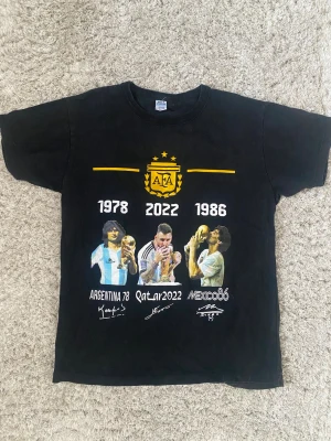Svart Argentina fotbollströja - Cool svart t-shirt med tryck som hyllar Argentinas VM-guld 1978, 1986 och 2022. Framsidan har AFA-logga, årtal och bilder på spelare med pokaler. Perfekt för fotbollsfans som vill visa sitt stöd för Argentina.