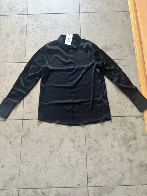 Svart satinblus från BikBok - Stilren svart skjorta från BikBok i glansigt satinliknande material. Klassisk krage, långa ärmar och knäppning framtill. Perfekt för dig som gillar en clean och elegant look.