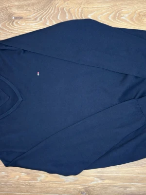 Mörkblå V-ringad tröja Tommy Hilfiger - Snygg mörkblå V-ringad tröja från Tommy Hilfiger med diskret logga på bröstet och rödvit detalj i kragen. Tillverkad i mjukt, tunt stickat material som passar perfekt till lager på lager. Klassisk och stilren design som funkar till det mesta.