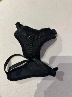 Svart sele för husdjur, Trixie - Säljer en svart sele för husdjur, tillverkad i slitstarkt meshmaterial med justerbara remmar och plastspännen. Selen har en metallring för koppel och är designad för att vara bekväm och säker vid promenader. Lite. Hund/katt