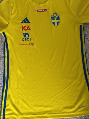 Svensk landslagströja Adidas gul S - Gul svensk landslagströja från Adidas i slim fit med blå detaljer och flera sponsorer som ICA, OBOS och Swedbank. Tröjan har SVFF-emblem på bröstet och är tillverkad i lätt, ventilerande material med AEROREADY-teknologi. Perfekt för fotboll eller träning.
