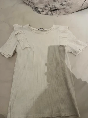 Vit ribbad topp med volangdetaljer - Supersöt vit ribbad topp med korta ärmar och volangdetaljer på axlarna. Perfekt till jeans eller kjol för en trendig look. Mjuk och skön känsla, enkel att matcha med olika stilar.