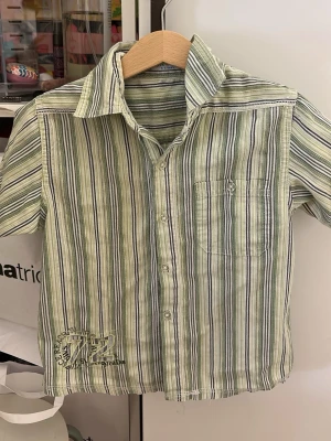 Randig grön skjorta med tryck - Kortärmad skjorta med gröna, vita och grå ränder. Skjortan har en bröstficka och ett broderat tryck med siffran 72 nedtill. Klassisk krage och knäppning framtill. Perfekt för dig som gillar en avslappnad men ändå stilren look.