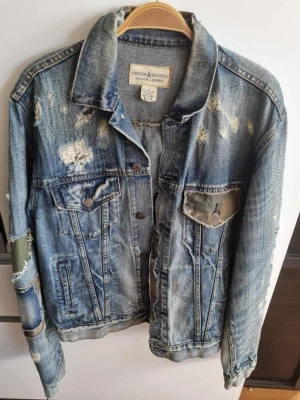 Distressed jeansjacka från Ralph Lauren - Säljer en blå jeansjacka med slitna detaljer och camo-tyg på ryggen och fickan. Jackan har klassisk krage, knappstängning och flera fickor fram. Rå och trendig look med patchade partier och trasiga effekter. Perfekt för dig som gillar streetstyle.